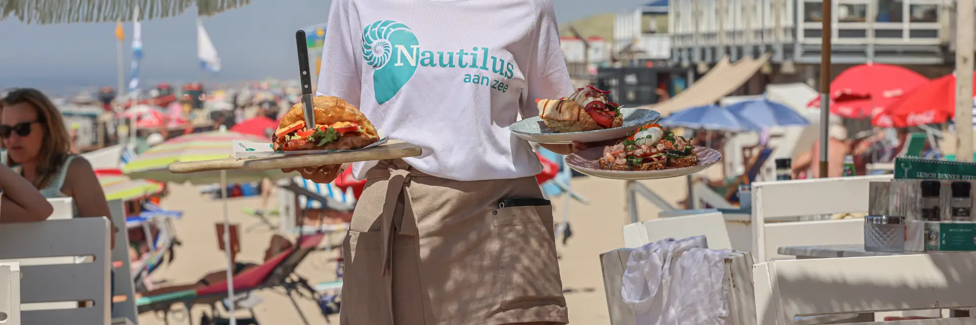 Medewerker in Egmond aan Zee serveert gerechten op een strandterras met uitzicht op zee, strand en kleurrijke parasols op de achtergrond.