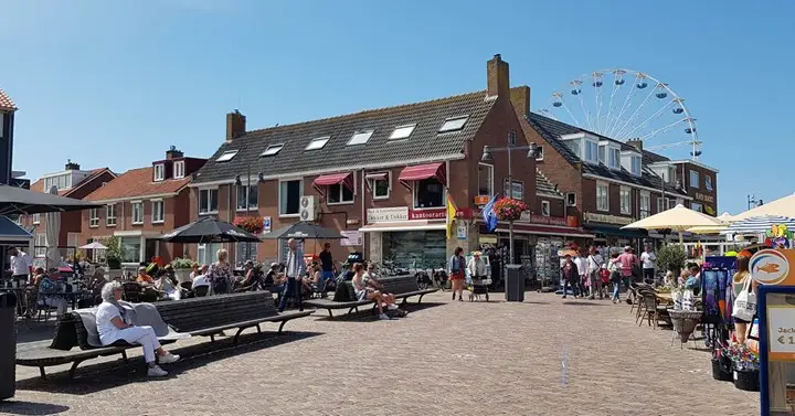 Foto van een bruisend Pompplein met mensen in het centrum van Egmond aan Zee waarop je een aantal winkels kunt zien zoals Boekhandel Dekker & Dekker. Met op de achtergrond ook het witte reuzenrad