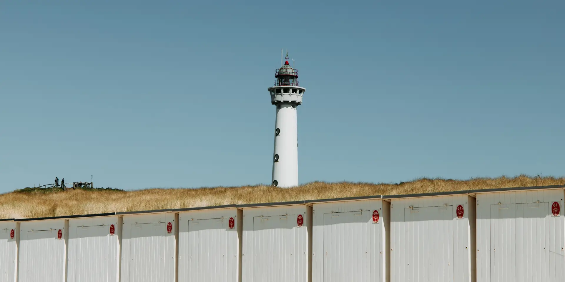 De vuurtoren van Egmond aan Zee, een witte vuurtoren steekt boven een duinlandschap uit, met een rij strandhuisjes op de voorgrond en een heldere blauwe lucht op de achtergrond