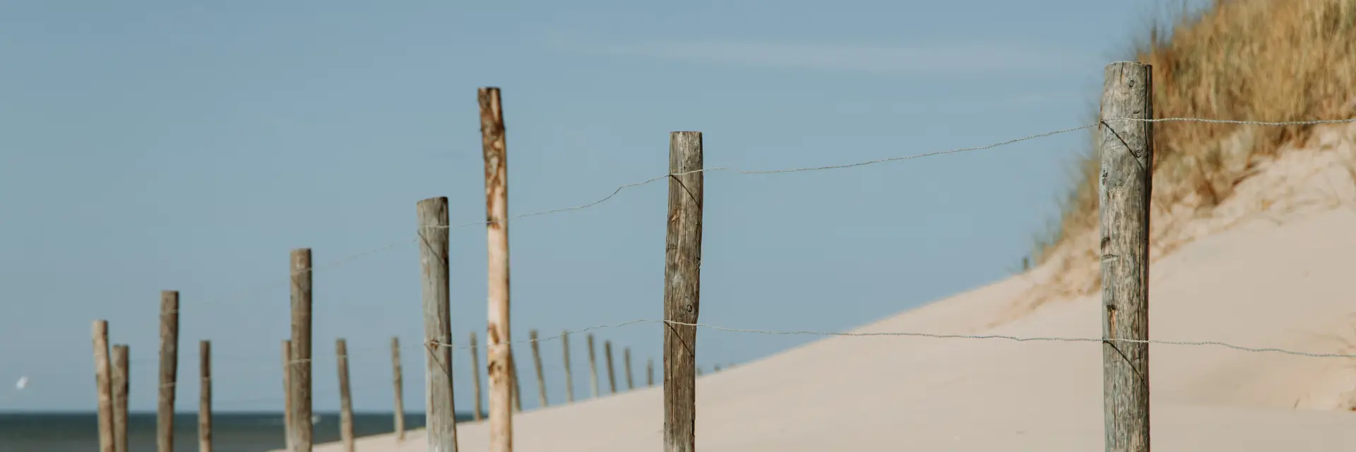 Een houten hekwerk met verweerde palen en gespannen draad loopt door een zandduin op het strand, met gras en de blauwe lucht op de achtergrond.