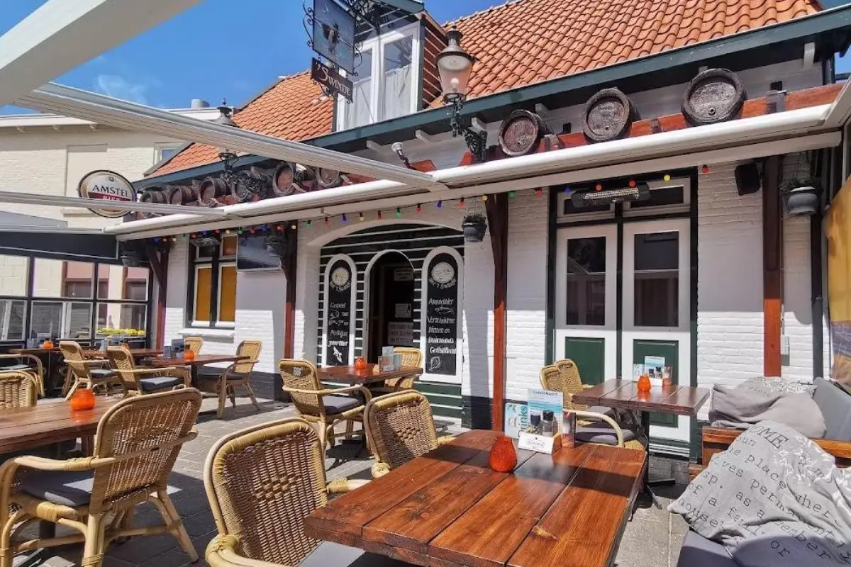 Een sfeervol terras van een café-restaurant met houten tafels en rotan stoelen, gelegen voor een traditioneel gebouw met een rood pannendak en witte gevel. Boven de ingang hangen decoratieve biervaten, en een uitschuifbaar zonnescherm.