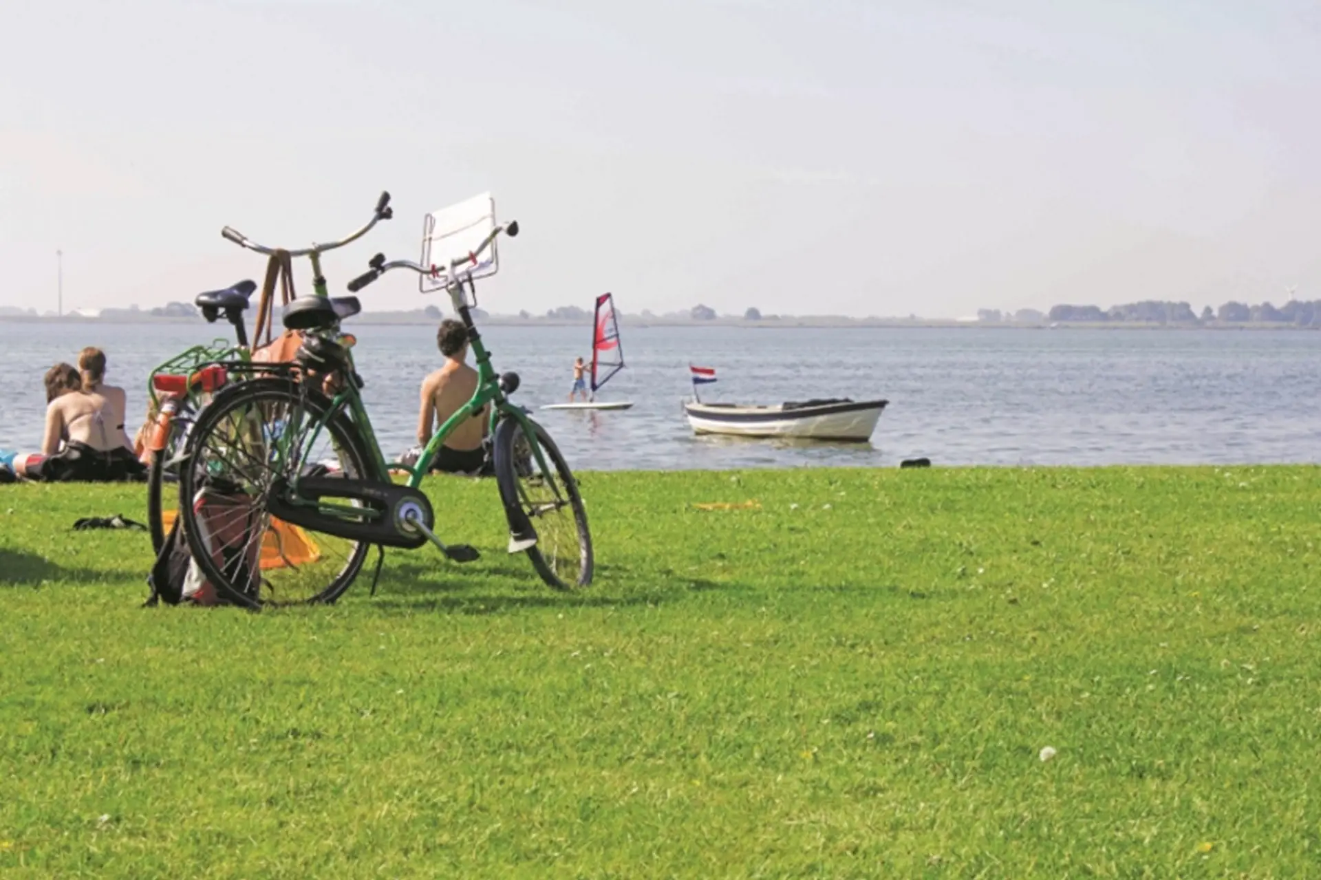 Mensen aan een meertje waar mensen aan het kitesurfen zijn en op de voorgrond staan twee fietsen.