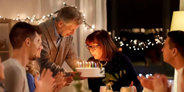Mevrouw blaast kaarsjes uit op een taart. Met allemaal mensen er omheen.