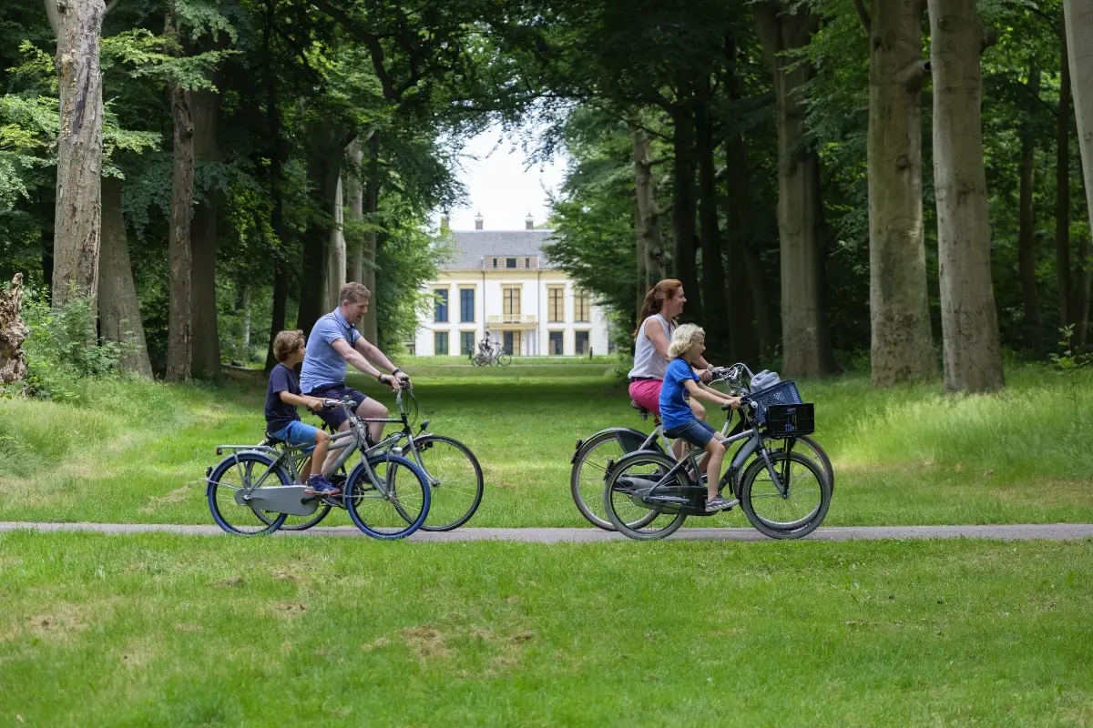 Een gezin fietst ontspannen over een pad door een groen park met hoge bomen. Op de achtergrond staat een statig wit landhuis. De kinderen zitten achterop bij hun ouders en genieten van de rit in de natuur
