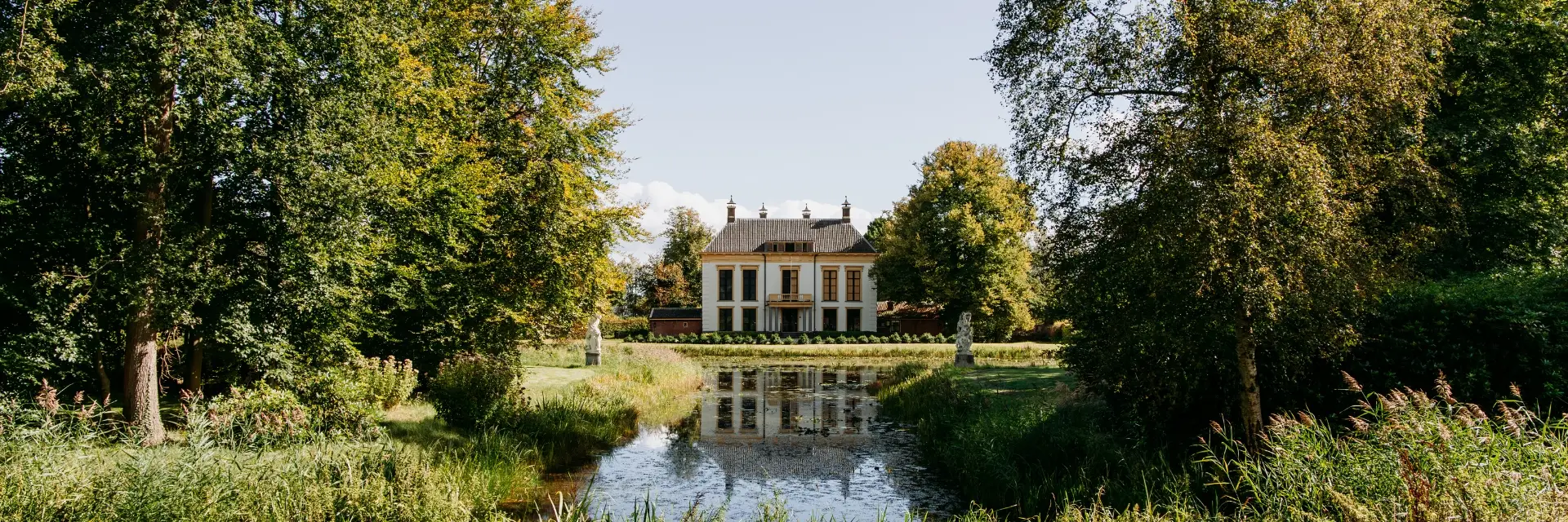Een statig, wit landhuis met een donker pannendak, gelegen te midden van een groene omgeving. Voor het huis ligt een vijver waarin het gebouw en de omliggende bomen reflecteren. Aan weerszijden van de vijver staan klassieke stenen beelden op sokkels.