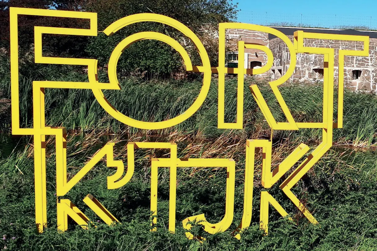 Een opvallend geel kunstwerk met de tekst "FORT K’IJK" staat voor het historische fort, dat deels zichtbaar is op de achtergrond. Het fort, met zijn karakteristieke stenen muren en kleine ramen, is omgeven door groen riet en natuurlijke begroeiing.