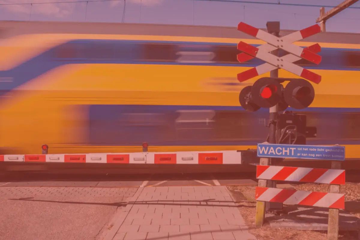 Een gele en blauwe trein rijdt met hoge snelheid voorbij een gesloten spoorwegovergang. De rode en witte slagbomen zijn naar beneden, en het waarschuwingsbord met de tekst 'WACHT' geeft aan dat oversteken nog niet veilig is