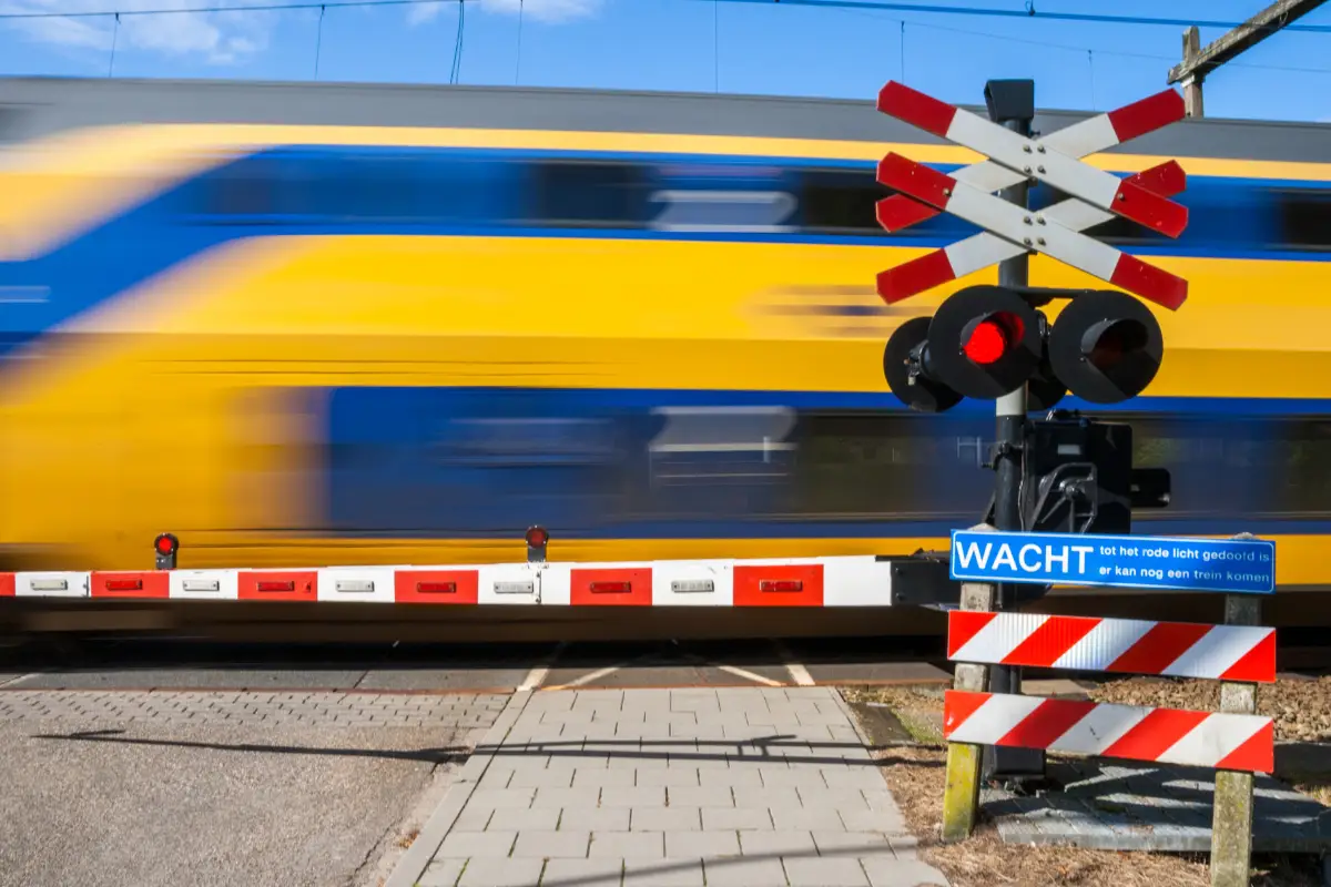 Een gele en blauwe trein rijdt met hoge snelheid voorbij een gesloten spoorwegovergang. De rode en witte slagbomen zijn naar beneden, en het waarschuwingsbord met de tekst 'WACHT' geeft aan dat oversteken nog niet veilig is