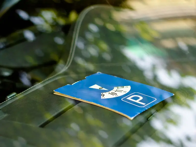Een blauwe parkeerschijf met een witte 'P' en een instelbare tijdsaanduiding ligt op het dashboard van een auto, zichtbaar door de voorruit. De reflectie van groene boombladeren geeft een zomerse sfeer aan het beeld.